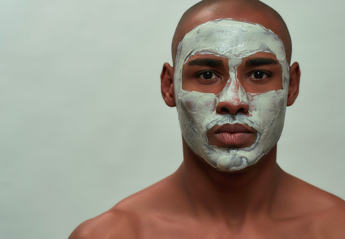 Portrait d'un homme de couleur sur fond vert pâle portant un masque exfoliant.